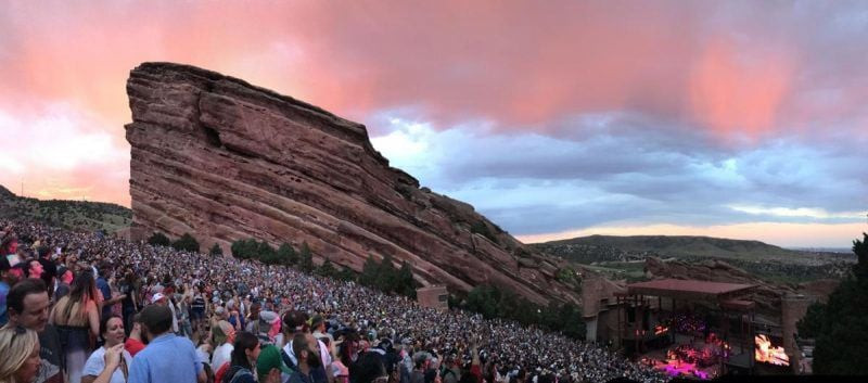 Red Rocks Amphitheatre during sunset, capturing the vibrant colors of the sky blending with the red rock formations and the stage lights illuminating the scene.