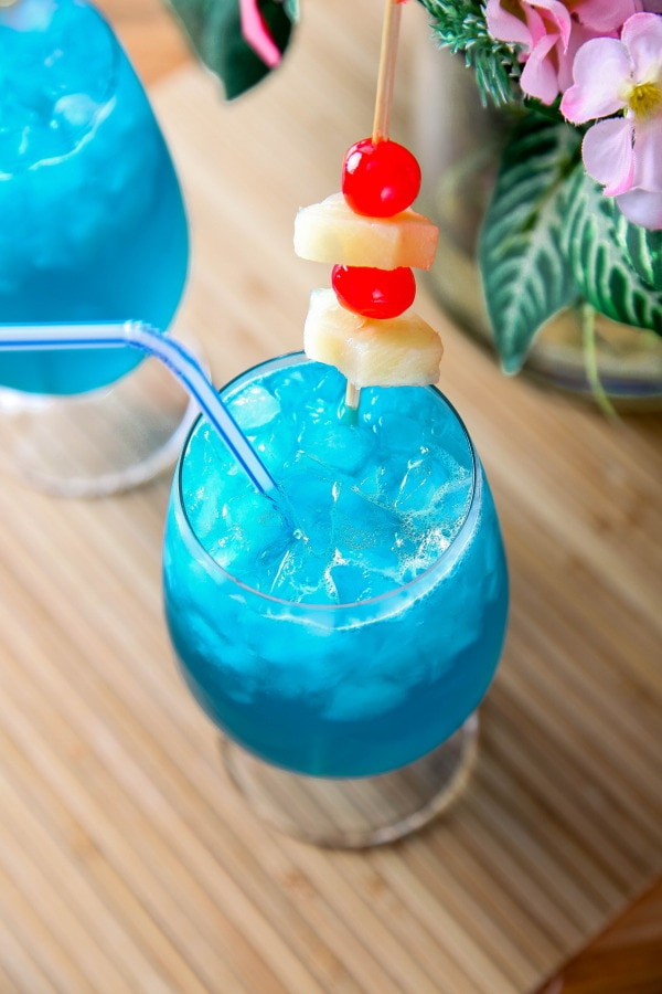 a closeup of a blue Hawaii cocktail garnished with pineapple and maraschino cherries.