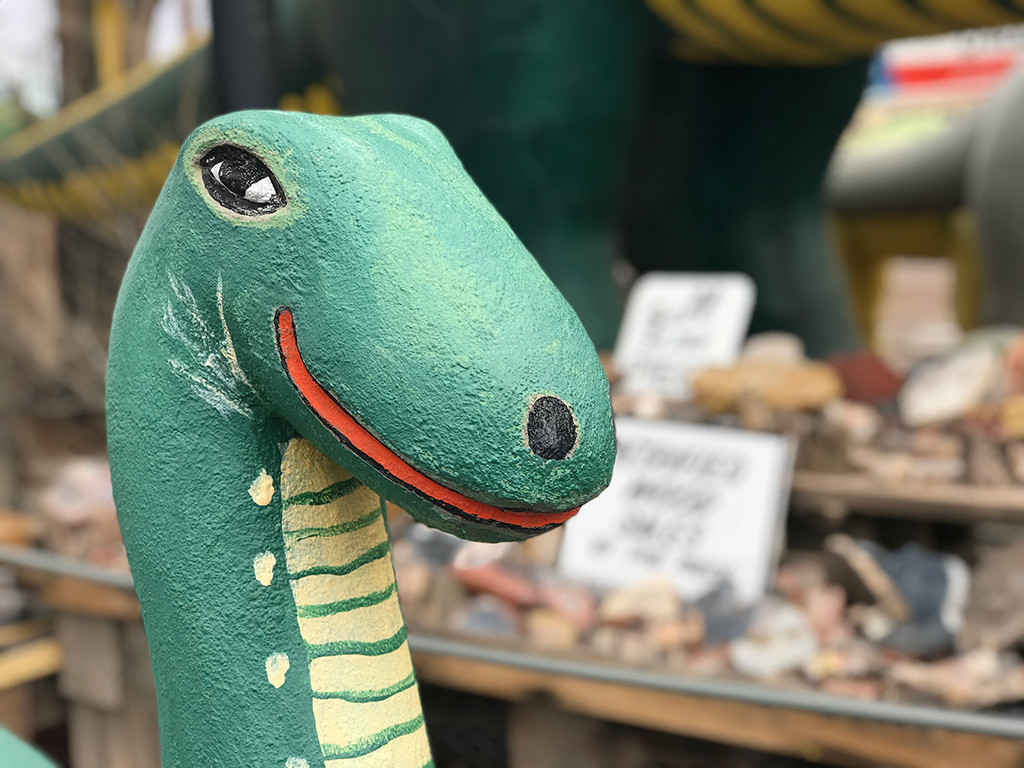 A close-up view of a large, green dinosaur statue at the Rainbow Rock Shop, with a clear blue sky in the background