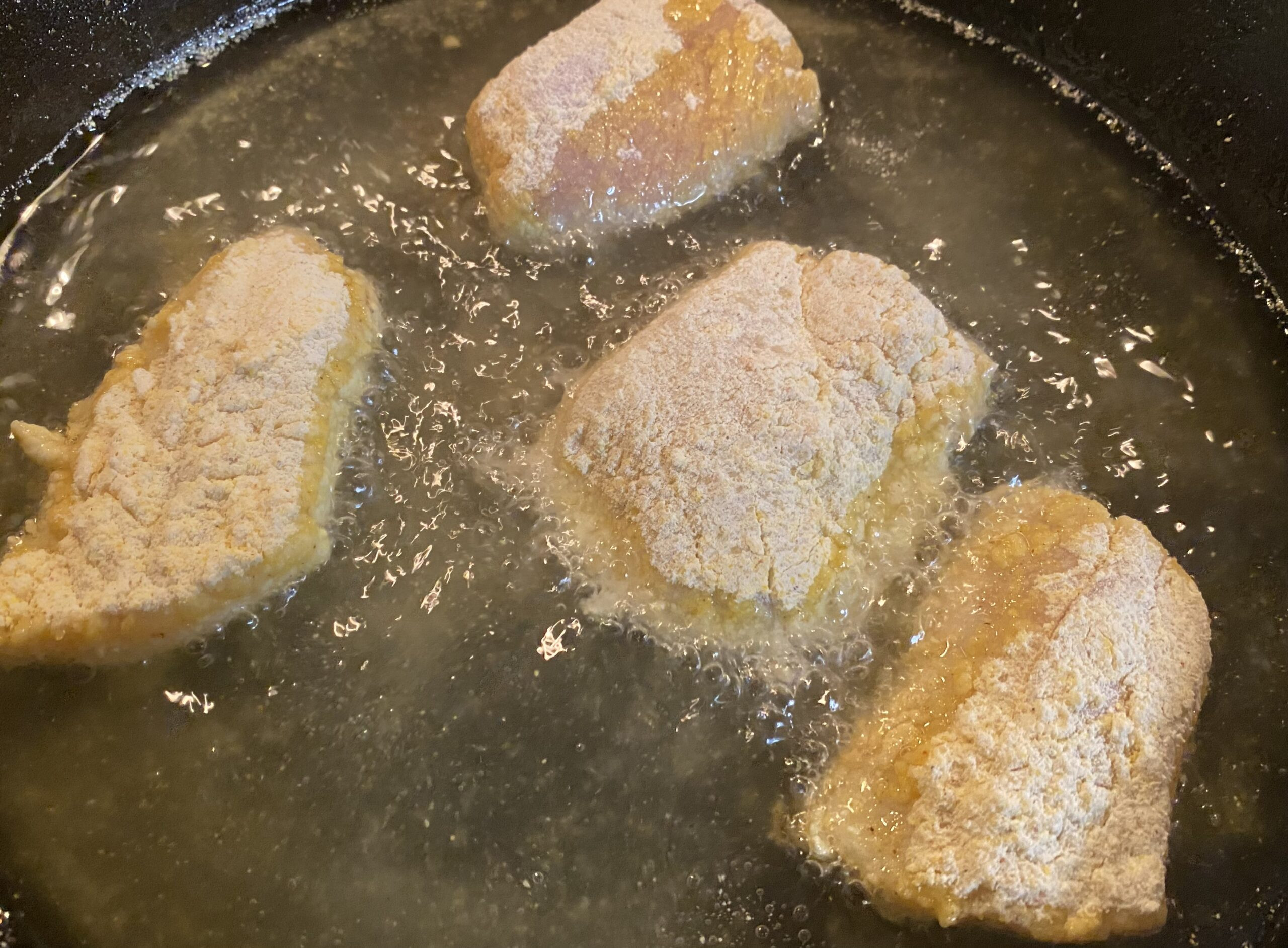 Rock fish fillets frying in hot oil in a cast iron skillet, turning golden brown.