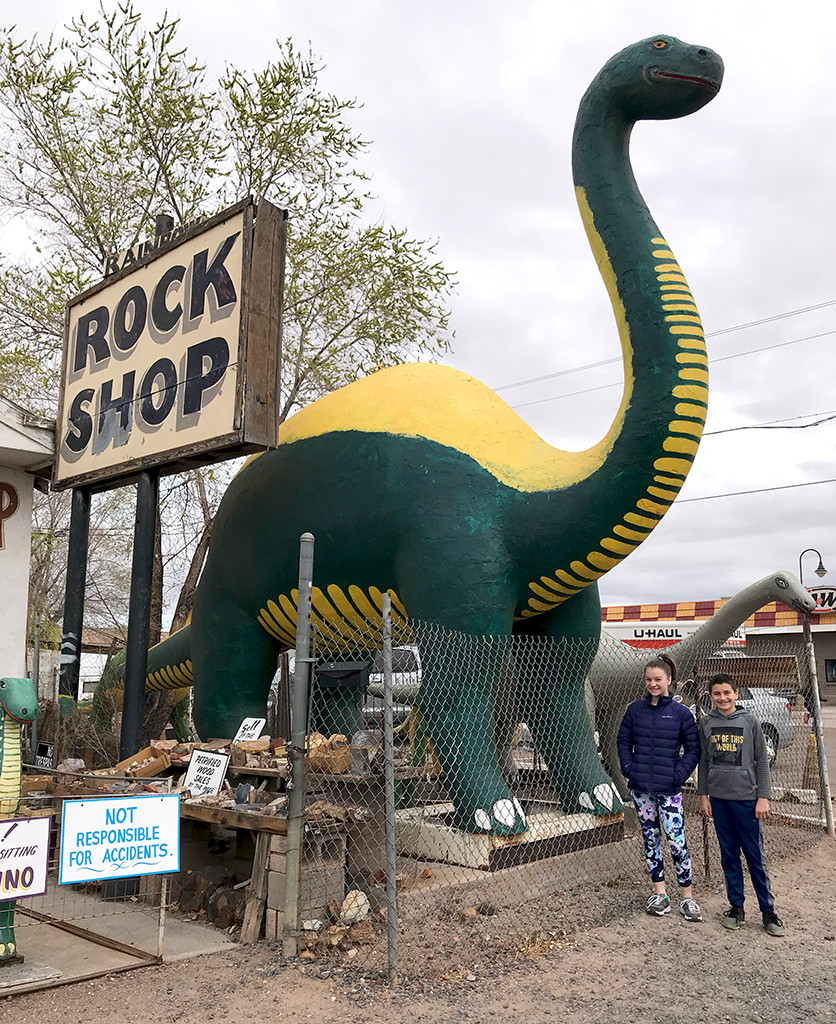 Two children, Natalie and Carter Bourn, posing with a blue dinosaur statue at the Rainbow Rock Shop, smiling at the camera