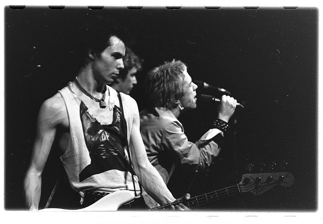 A black and white photo captures the raw energy of a live punk rock performance, with a band intensely playing instruments on stage, bathed in dramatic lighting and a backdrop of amplifiers, evoking the rebellious spirit of punk rock.