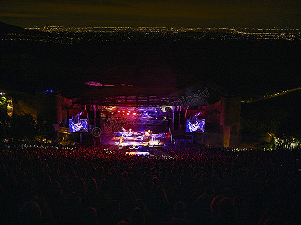 Panoramic view from the top row of Red Rocks Amphitheater during a Lord Huron concert, city lights of Denver visible in the distance.