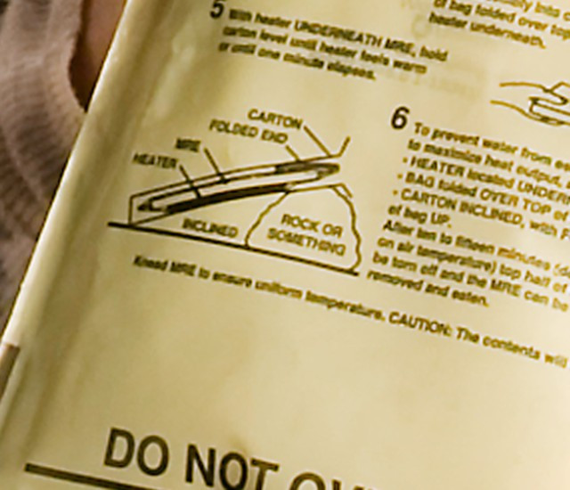 Close-up of a Flameless Ration Heater (FRH) package, highlighting the iconic phrase "rock or something" in the instructions. The FRH is placed at an angle with a small rock underneath, illustrating the original context of the phrase.