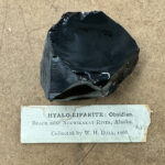 Black obsidian rock, shiny and reflective, displayed on a textured fiberboard surface with a vintage museum label.