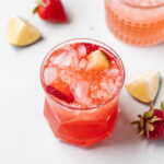 A clear, short glass with crushed ice, showcasing a strawberry margarita, surrounded by fresh limes and strawberries, embodying a cool, refreshing ambiance.