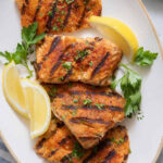 Plater of 4 grilled cod filets garnished with lemon wedges and chopped parsley
