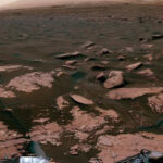 A panoramic view of the Martian surface captured by a rover. The foreground shows rocky terrain with various sized rocks and layered geological formations, indicative of past environmental conditions. The middle ground displays a smoother surface leading to a horizon that meets a hazy, dusty sky. Parts of the rover’s hardware are visible at the bottom edges of the image, confirming the image is taken by the rover itself.
