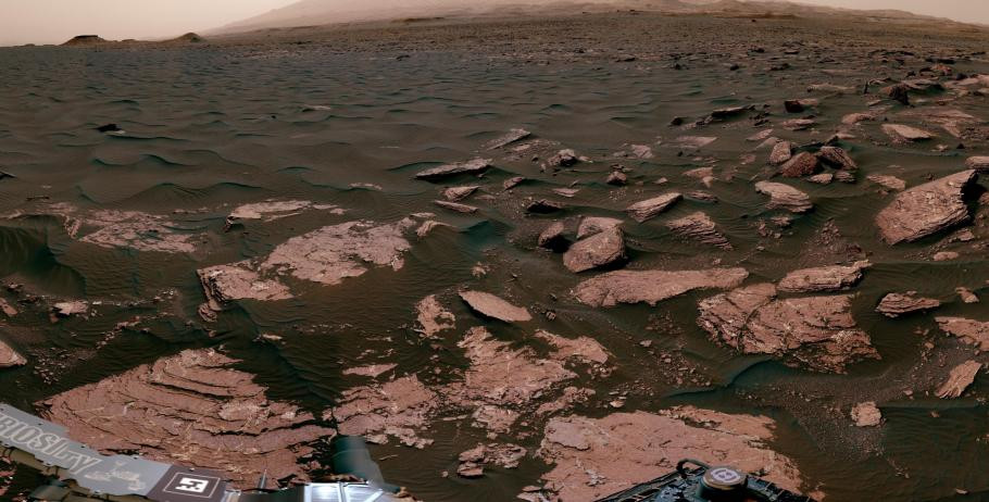 A panoramic view of the Martian surface captured by a rover. The foreground shows rocky terrain with various sized rocks and layered geological formations, indicative of past environmental conditions. The middle ground displays a smoother surface leading to a horizon that meets a hazy, dusty sky. Parts of the rover’s hardware are visible at the bottom edges of the image, confirming the image is taken by the rover itself.