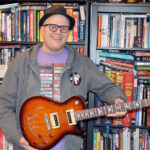 Adam Reader in his office holding an electric guitar given to him by Neal Schon, showcasing his passion for rock music and personal connections with iconic musicians.