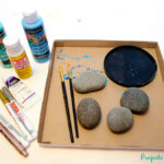 Supplies for drawing mermaid scales on painted rocks, including rocks, paintbrushes, and acrylic paints.