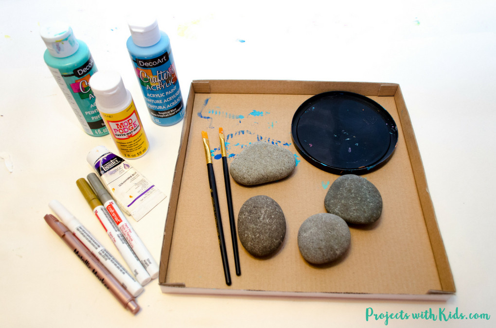 Supplies for drawing mermaid scales on painted rocks, including rocks, paintbrushes, and acrylic paints.