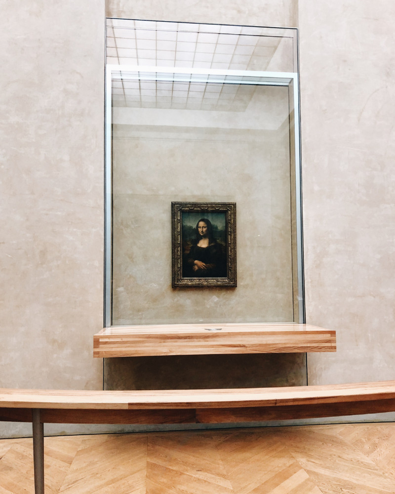 A photo of the Mona Lisa hanging in the Louvre behind a barrier and bulletproof glass case